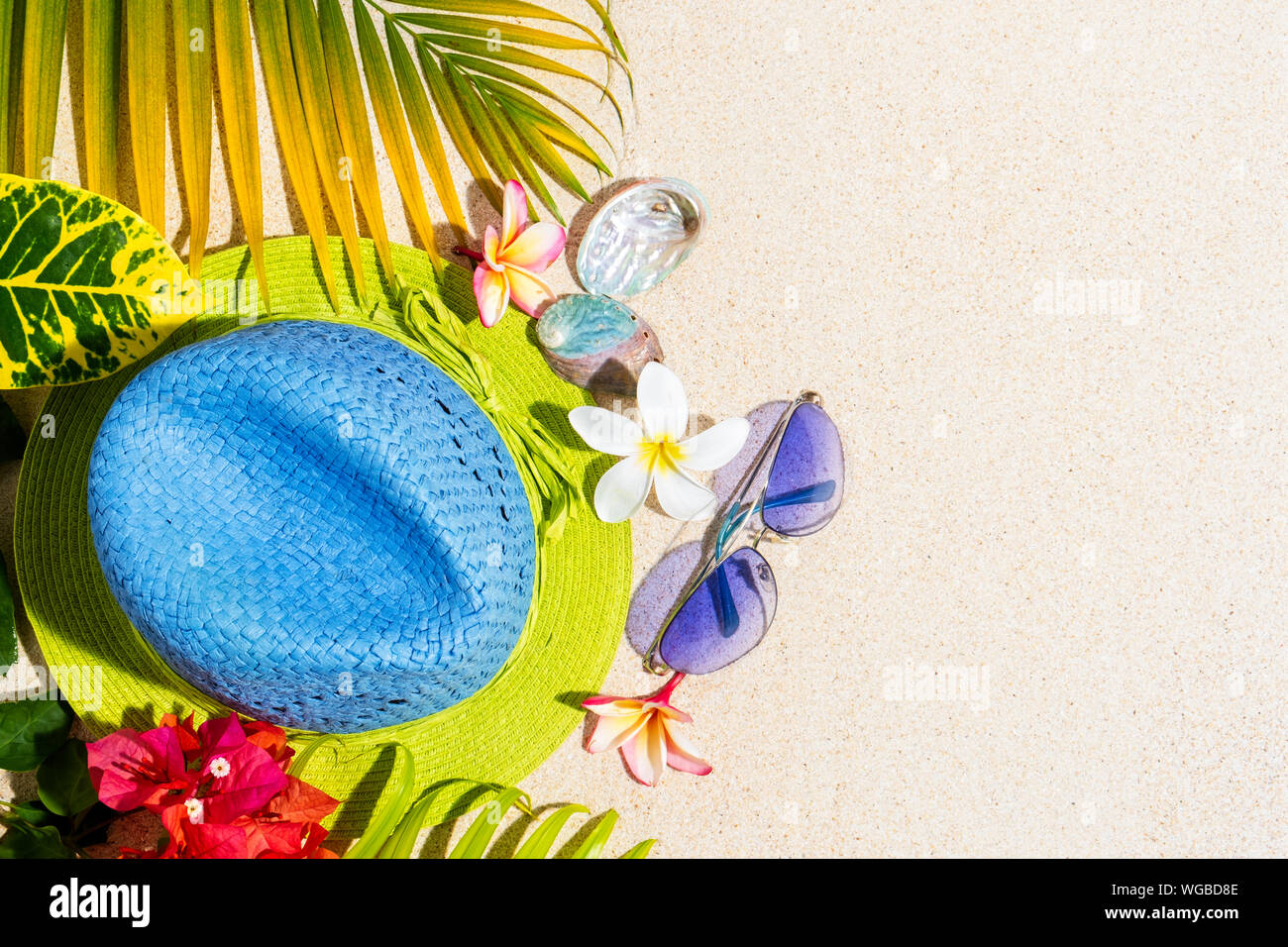 Blue and green straw hat with sunglasses, sea shells and frangipani flowers with green palm leafs on sand, summer vacation concept, top view, copy sap Stock Photo