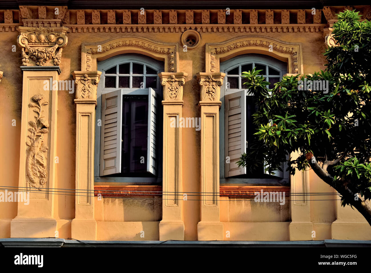 Front view of Singapore shop house with ornate exterior and grey wooden shutters flanked by tree in historic Geylang, Singapore Stock Photo