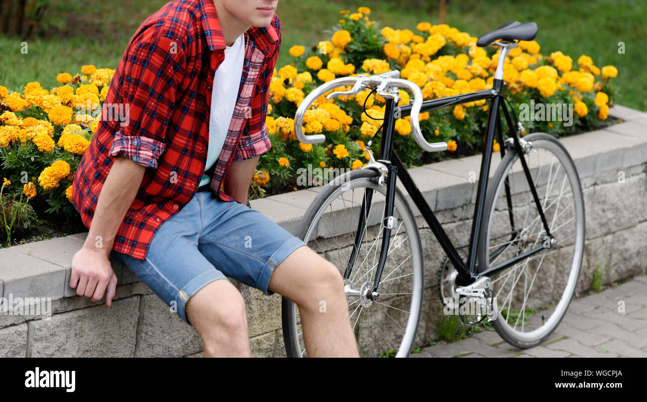 Hipster man with bicycle resting over flowerbed withal yellow flowers. WIthout face. Side view Stock Photo