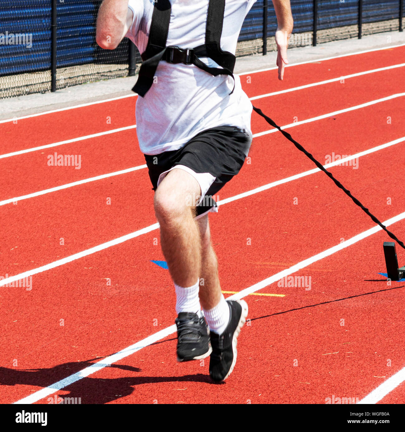 A high school male track and field athlete is pulling a sled with weights on a red track during