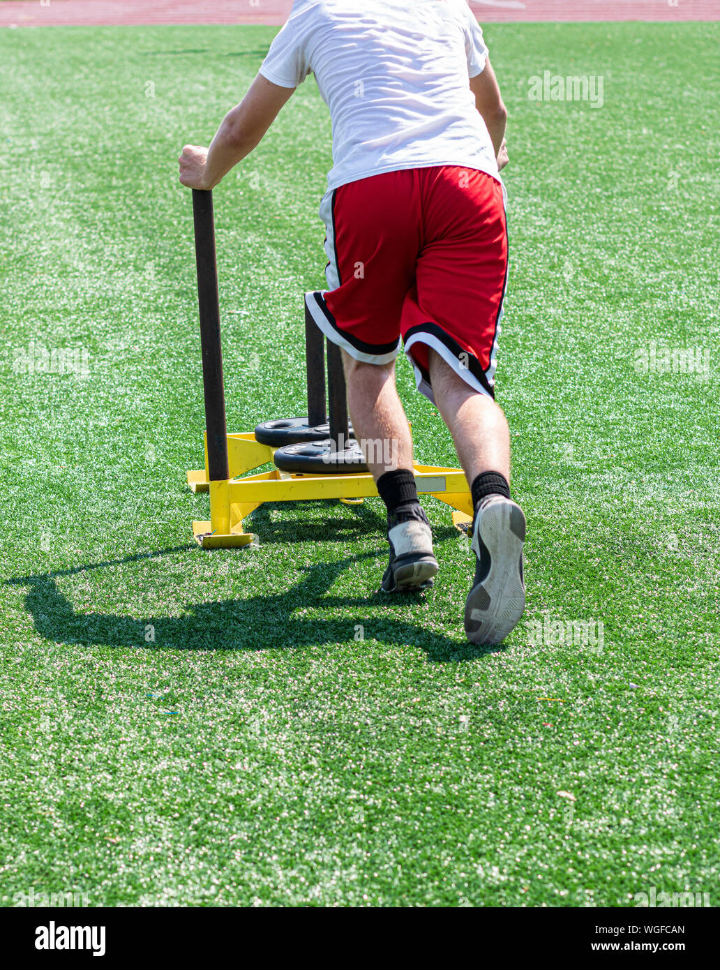A teenage boy is pushing a yellow sled with weights on it during running camp on a green turf field. Stock Photo