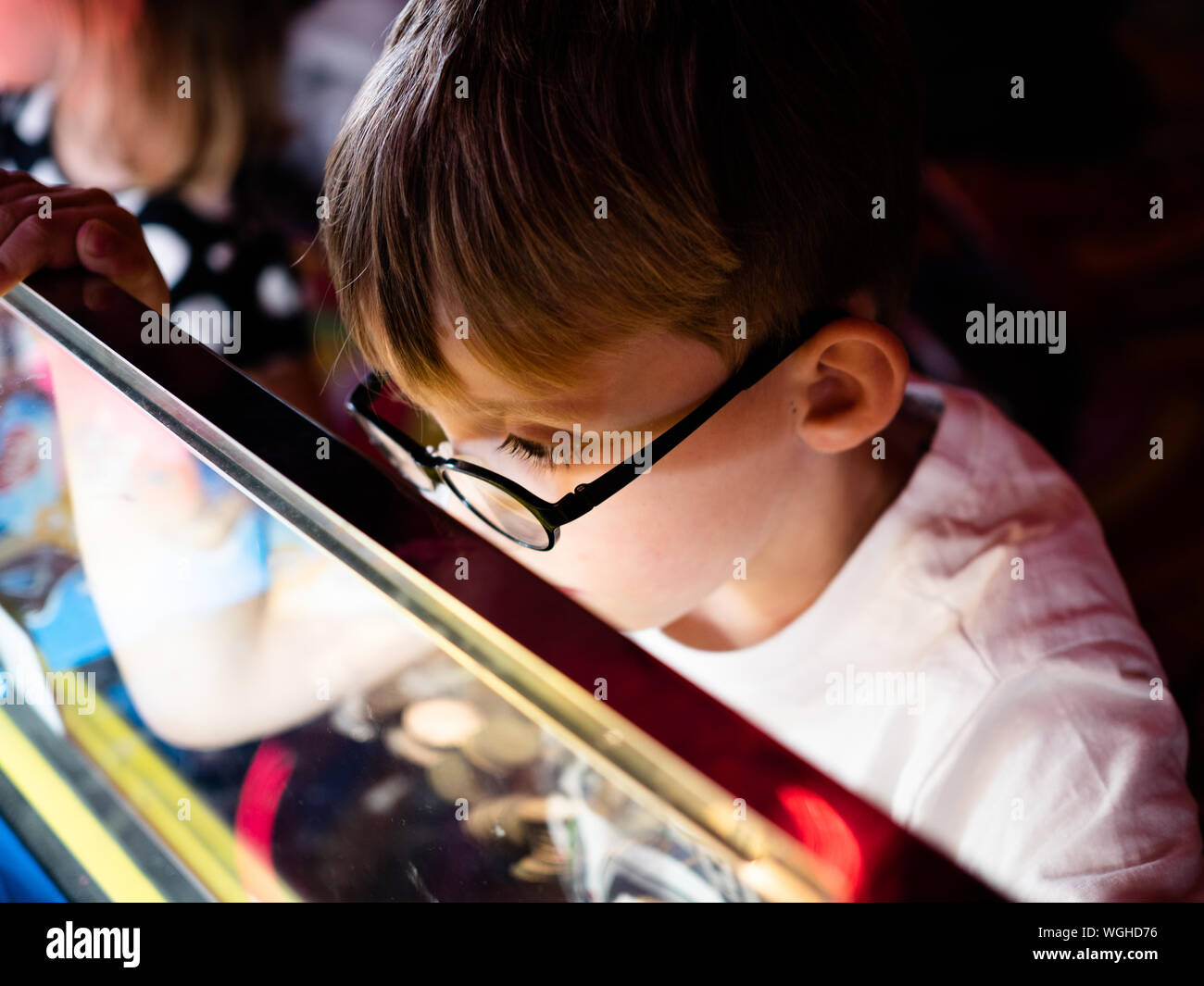 A 5-year old blonde boy wearing glasses watches intently whilst playing a 2p coin slot machine in a games arcade at a British seaside town. Stock Photo