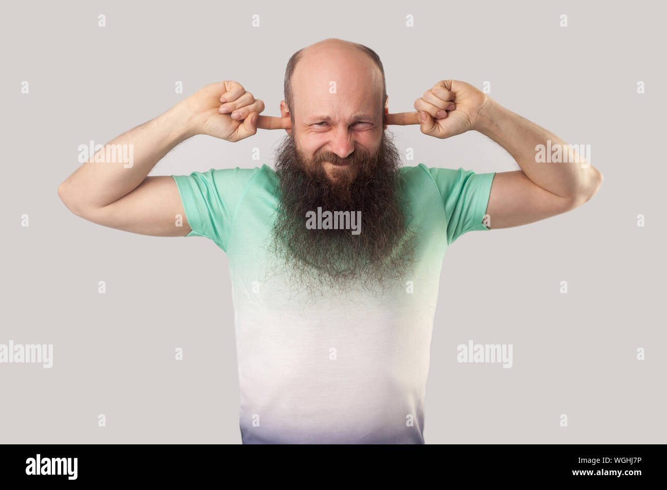 I don't want to hear you. Portrait of confused middle aged bald man with long beard and t-shirt standing with finger on his ears and blocking sound. i Stock Photo