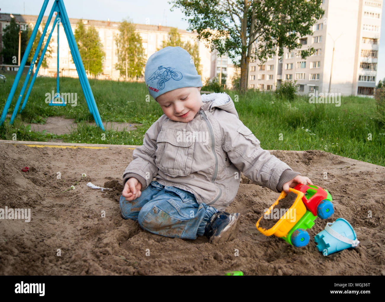 Russia, Novodvinsk - July 2019. A fair-haired boy of three years in a blue hat and jeans, in a light jacket plays in the sandbox with toys Stock Photo