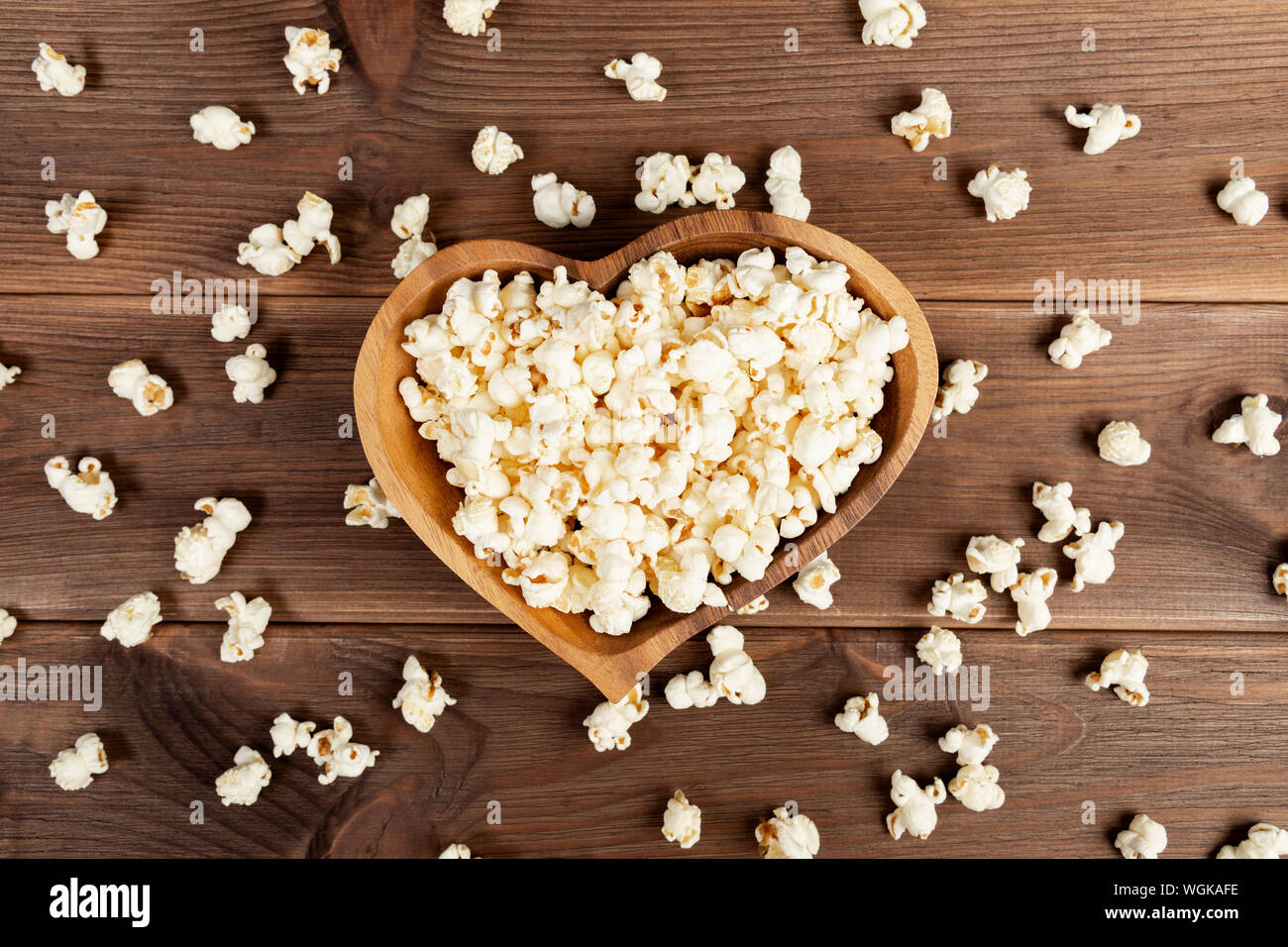 Popcorn in a brown wooden plate in the shape of a heart on a wooden table. Popcorn is laid out on the table Stock Photo