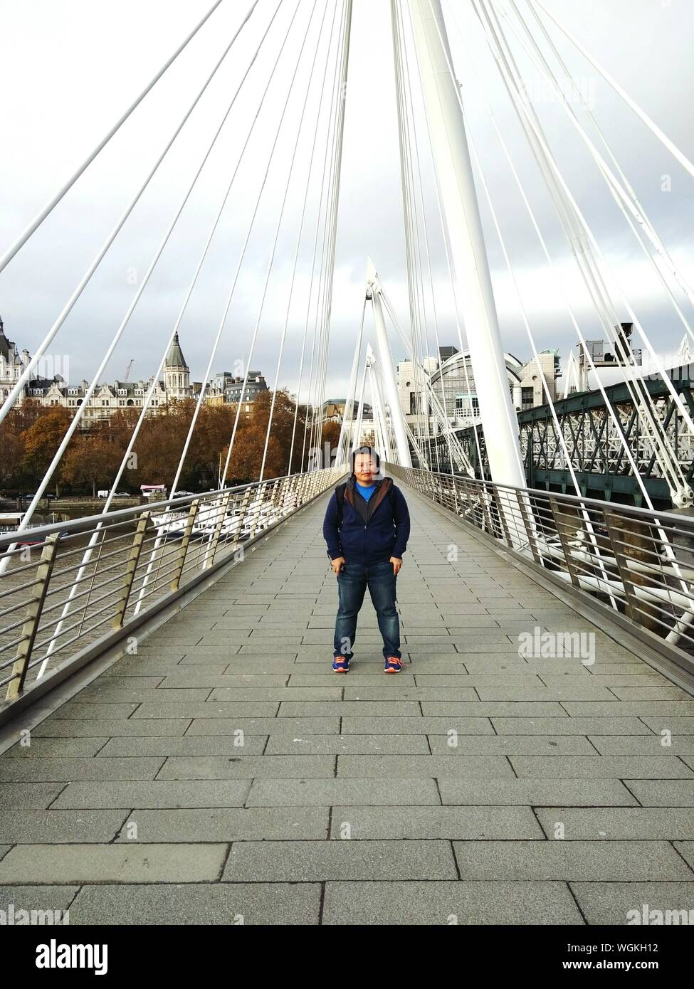 Full Length Of Man Standing On Golden Jubilee Bridge Against Cloudy Sky Stock Photo
