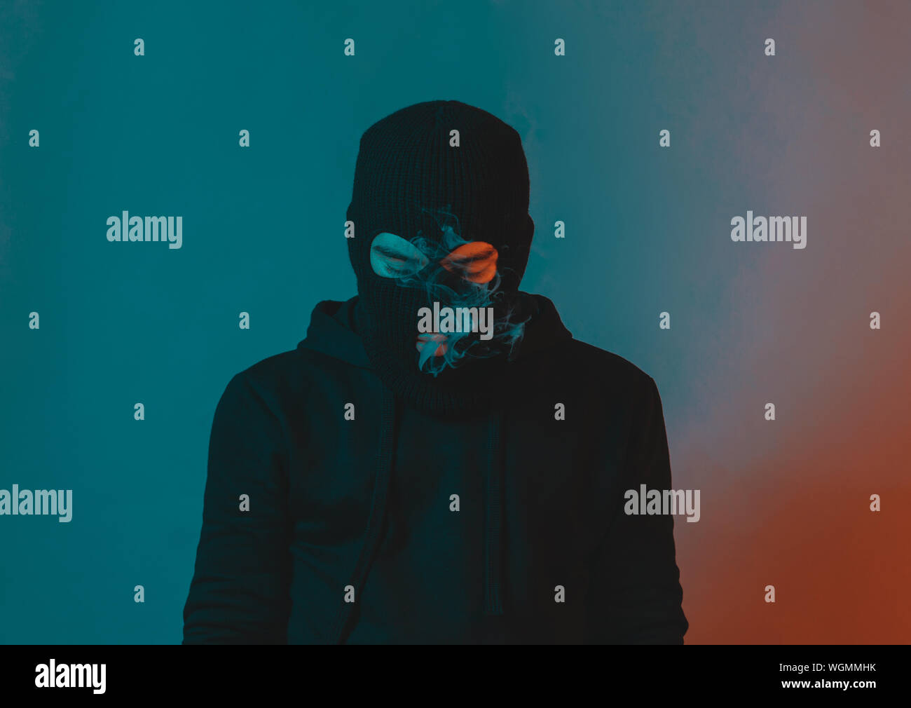 young man in hoodie and balaclava looking down and blow smoke out Stock Photo