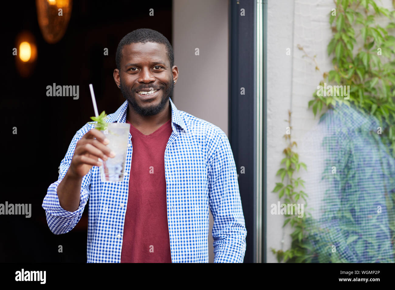 Waist up portrait of smiling African-American man holding cold drink outdoors while posing by cafe, copy space Stock Photo