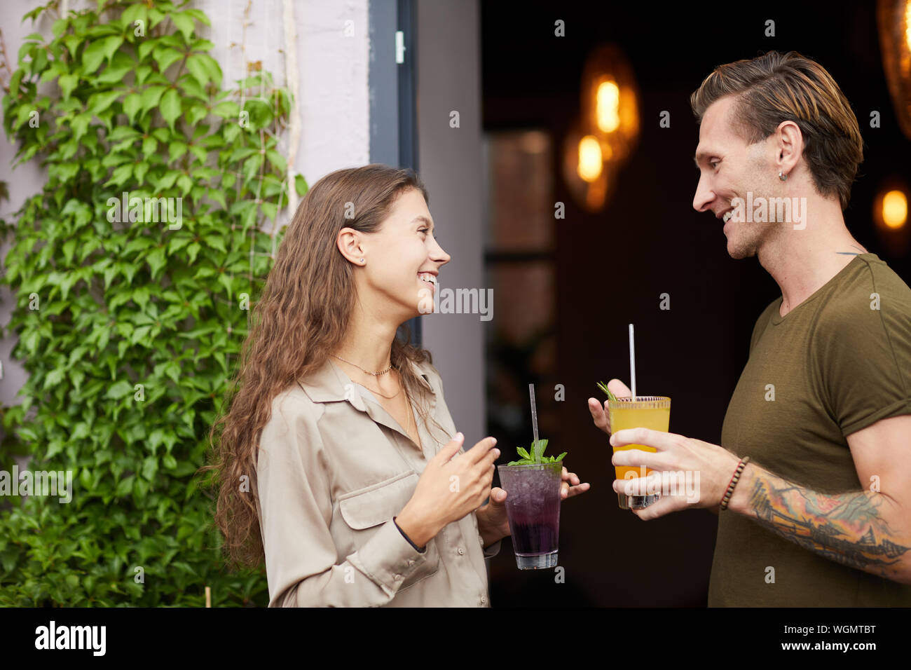 Side view portrait of smiling young woman talking to man outdoors standing by cafe door, both holding cold refreshing drinks, copy space Stock Photo
