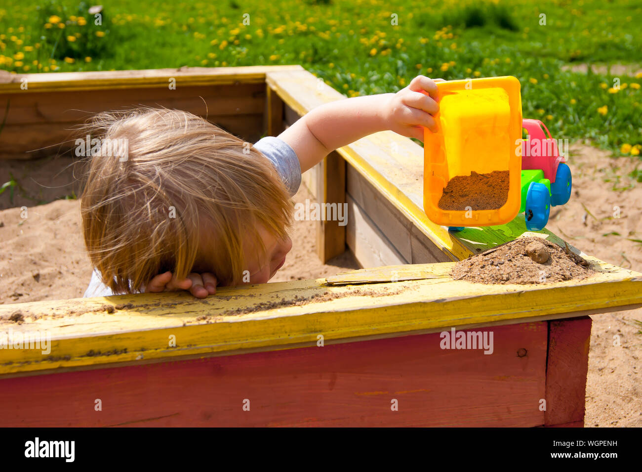 Summer in a small provincial town. Heat. A little blond boy of three years old tiredly lay down on a sandbox board, playing with his machine in the sand Stock Photo