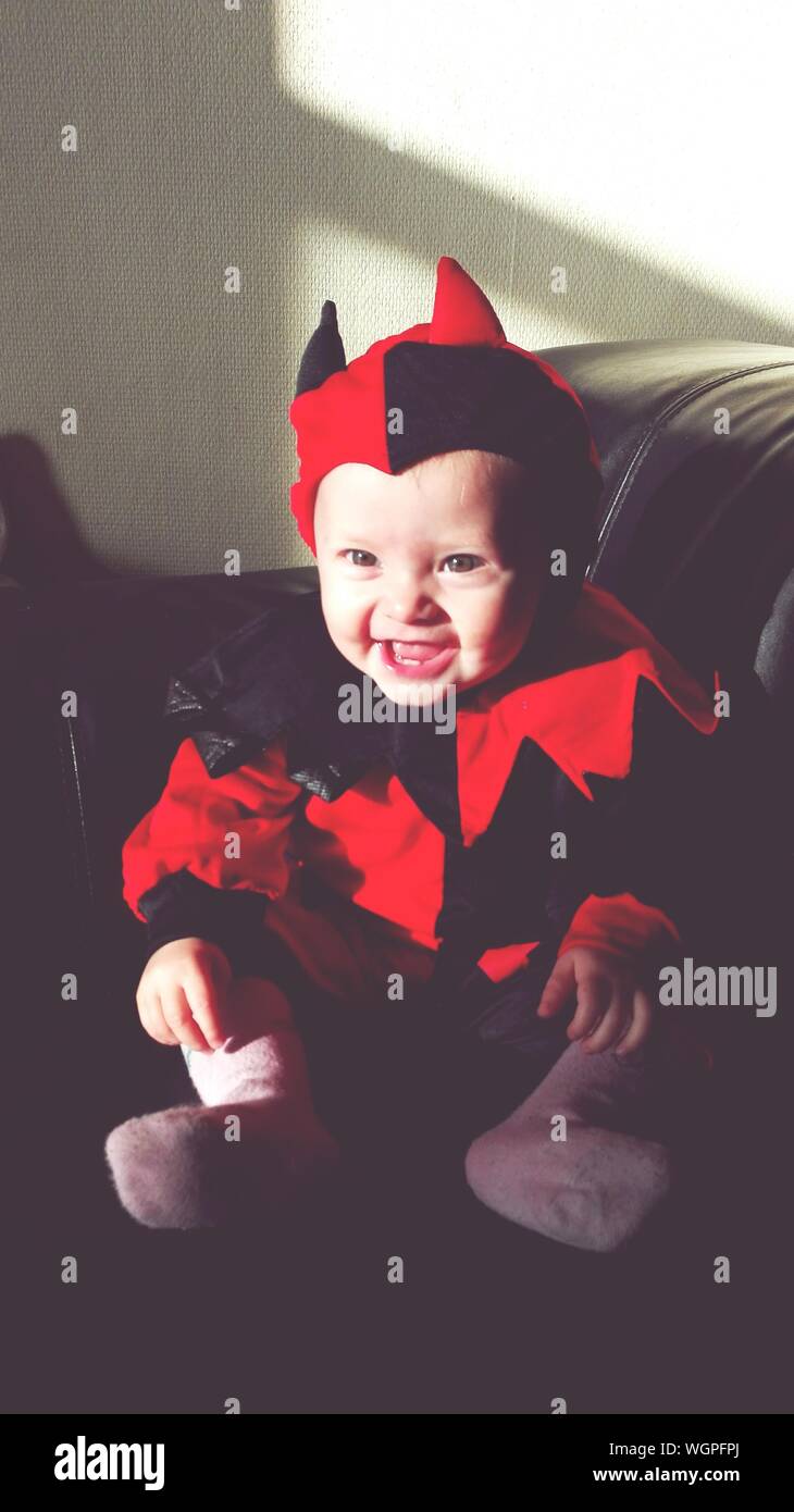 Cute Cheerful Baby Girl Wearing Red Devil Costume Sitting On Sofa At Home  Stock Photo - Alamy