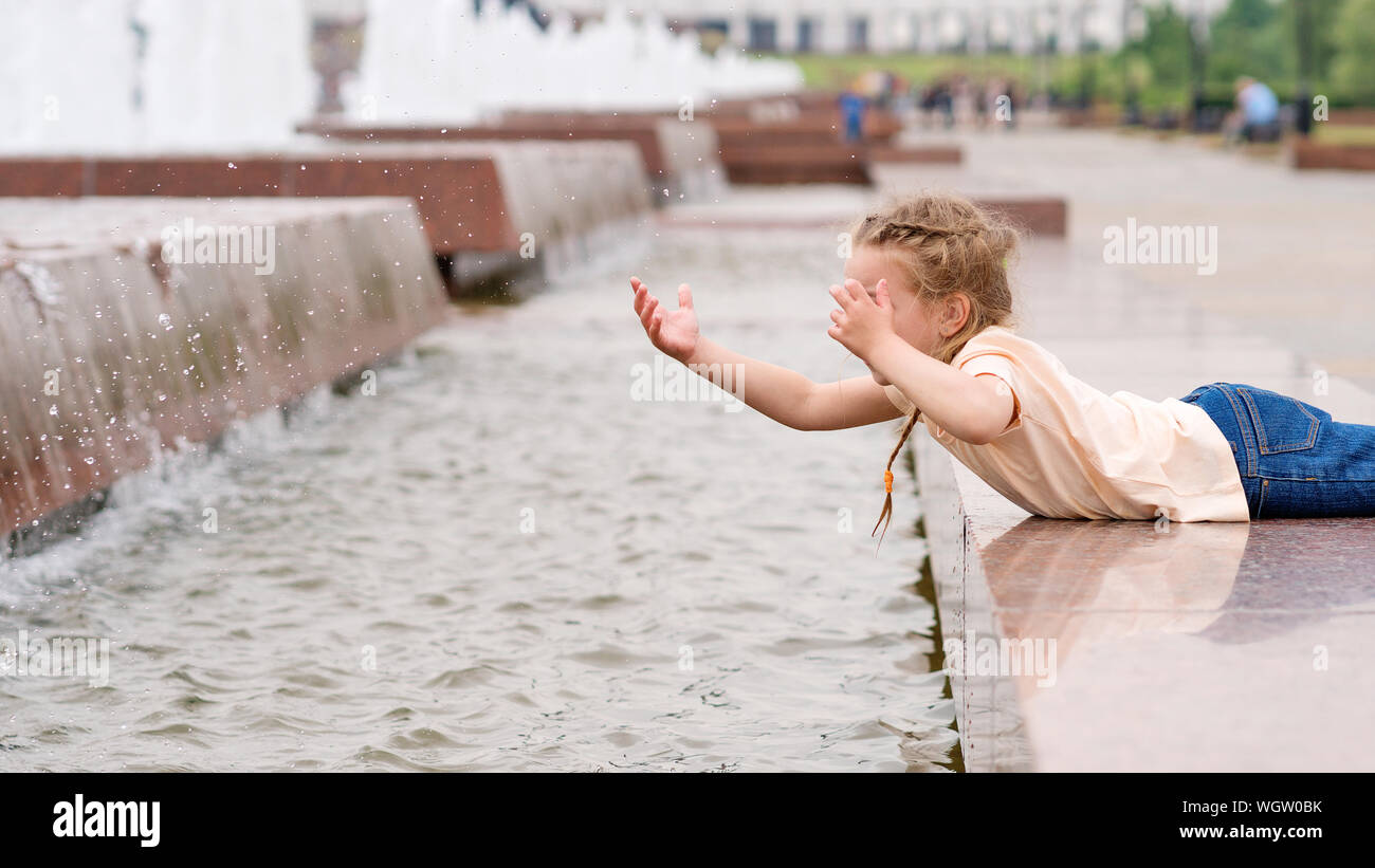 Сute little mischievous girl plays at the fountain. Time of prank. Urban casual outfit. Carefree. Happiness, fun and childhood concept. Stock Photo