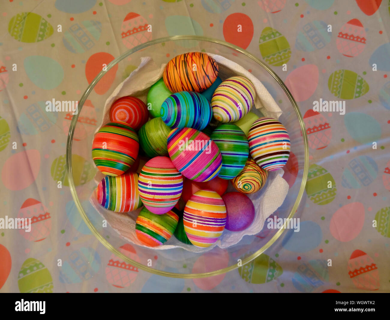 A collection of eggs dyed for easter in a glass bowl which is sitting on a plastic table cloth imprinted with an easter theme. Stock Photo