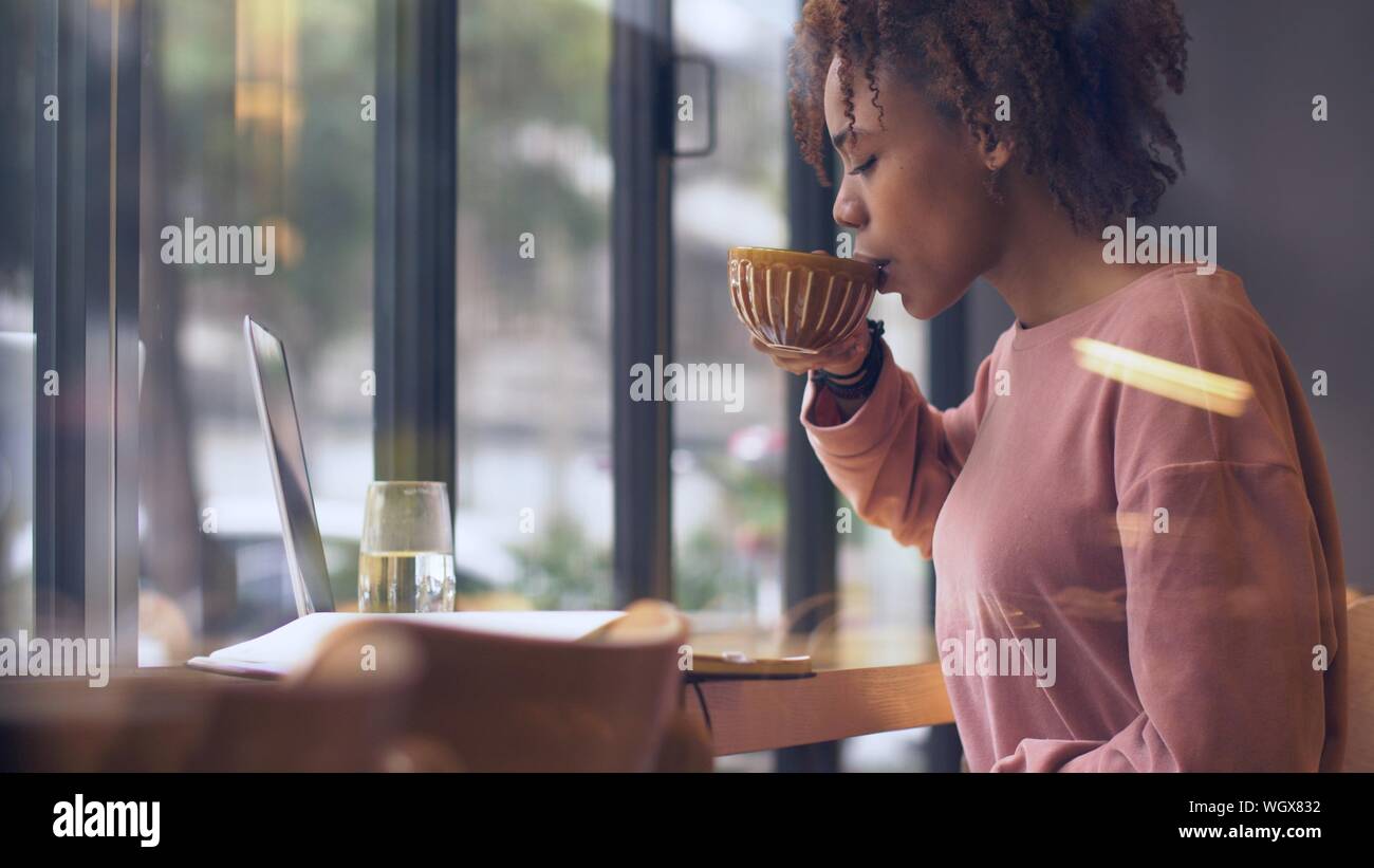 Side view of pretty young African woman drinking coffee indoor working ...