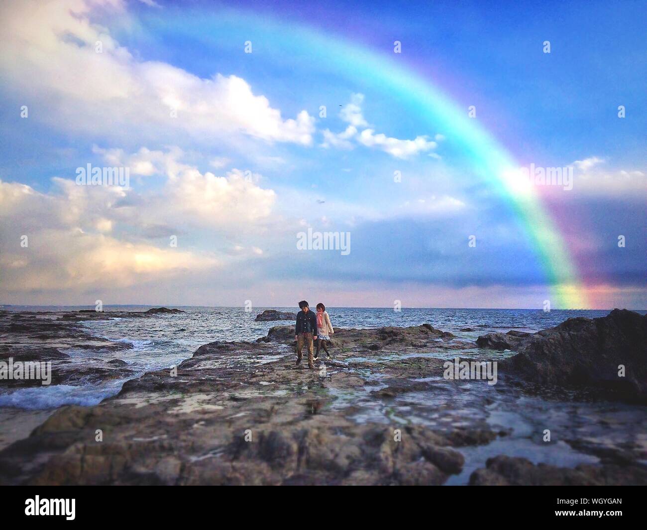 Young Adults Walking On Rocks Against Rainbow Sky Stock Photo