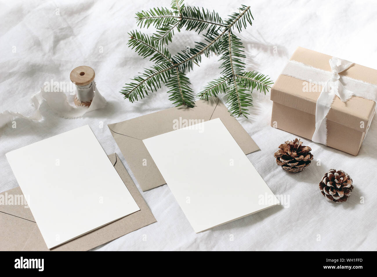 Christmas table, festive styled composition. Christmas greeting cards, envelopes mock-ups. Handmade gift box with silk ribbon, pine cones and fir tree Stock Photo