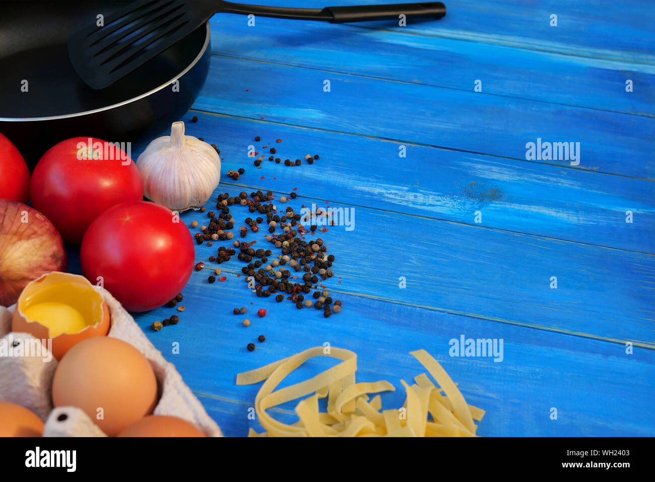 Breakfast Set. Frying pan, tomatoes, raw eggs, onions, garlic, spices, dry pasta. Selective focus. Copy space. On blue wooden background. Close-up. Stock Photo