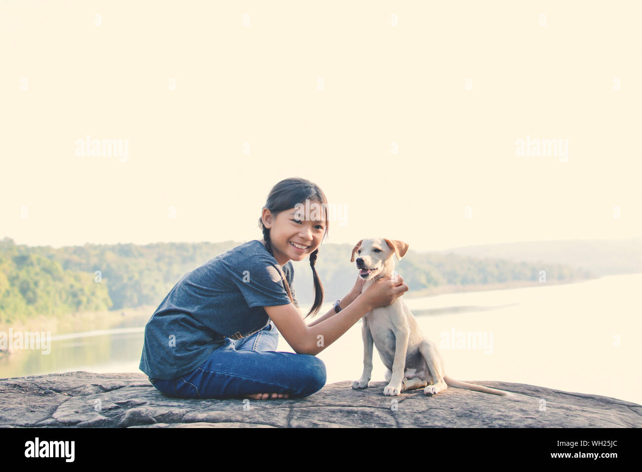 Side View Of Happy Girl Sitting With Puppy On Rock Against Lake Stock Photo