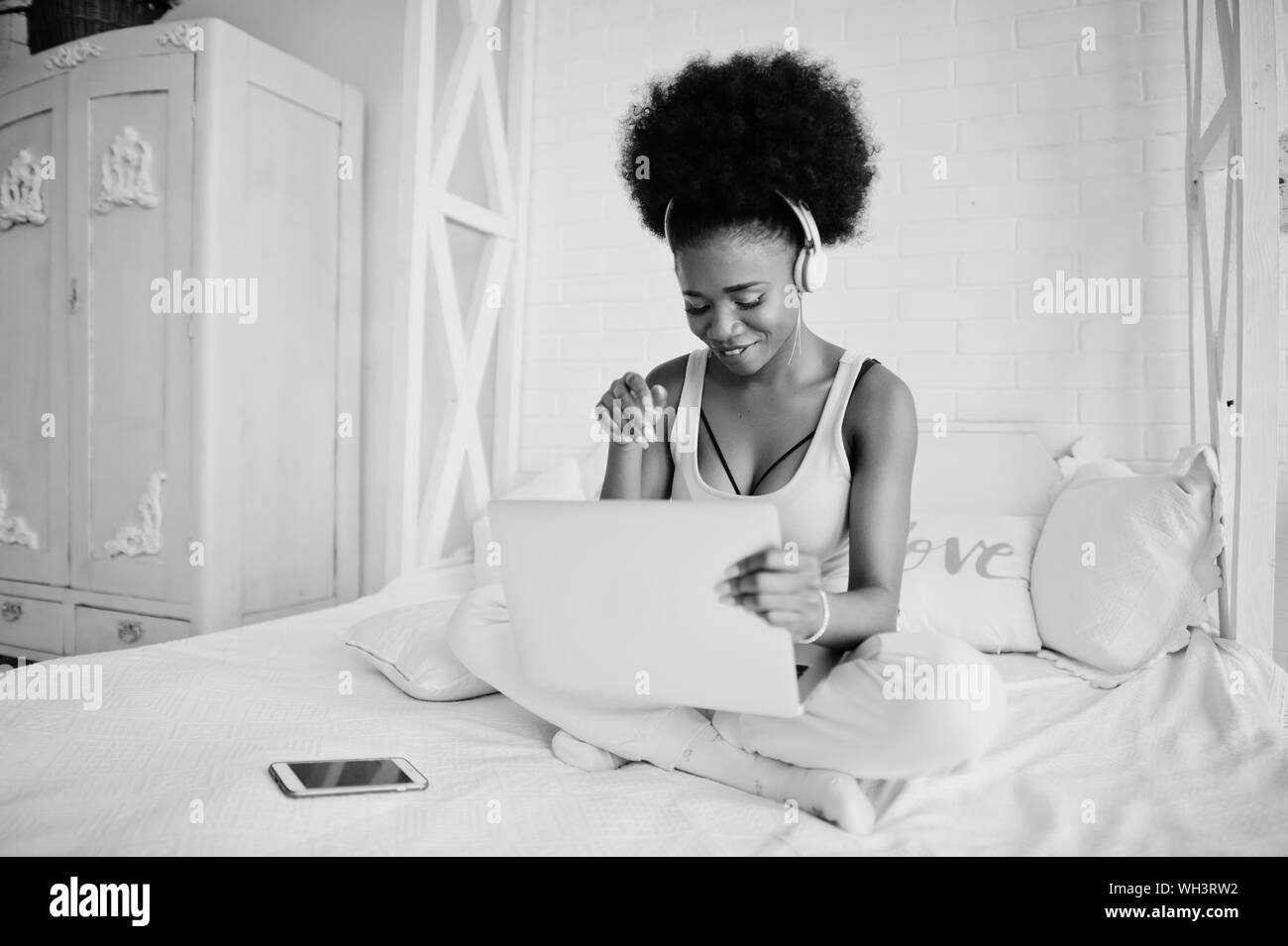 Young african american woman sitting in bed while working on laptop and listen music on earphones. Stock Photo