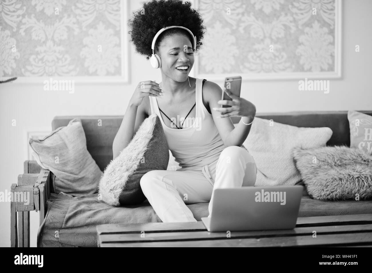 Young african american woman sitting on the couch while working on laptop and listen music on earphones, streaming video on her phone. Stock Photo