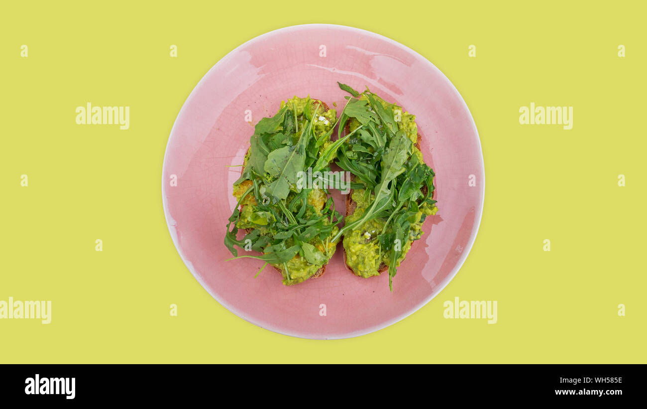 Two slices of mashed avocado toast garnished with fresh rocket or arugula salad set on a pink plate against a lime green background Stock Photo