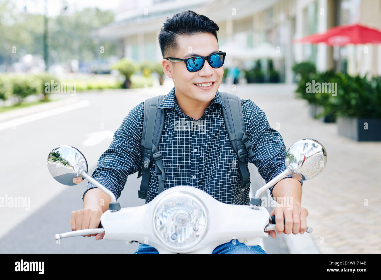 Happy young smiling Vietnamese man with backpack enjoying riding in city streets on scooter Stock Photo