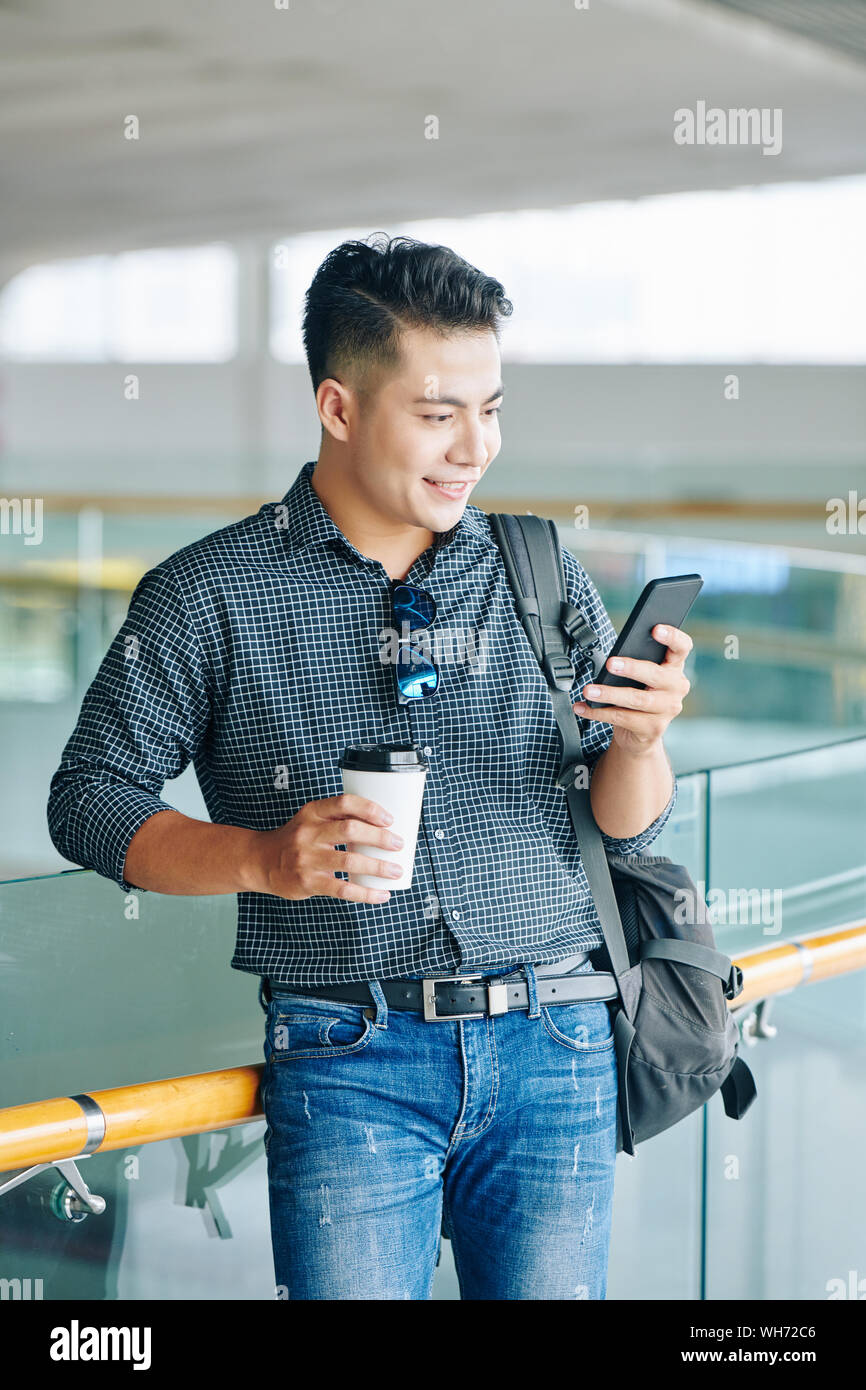 Smiling young Vietnamese man with backpack standing in airport terminal, drinking coffee and checking schedule in smartphone Stock Photo