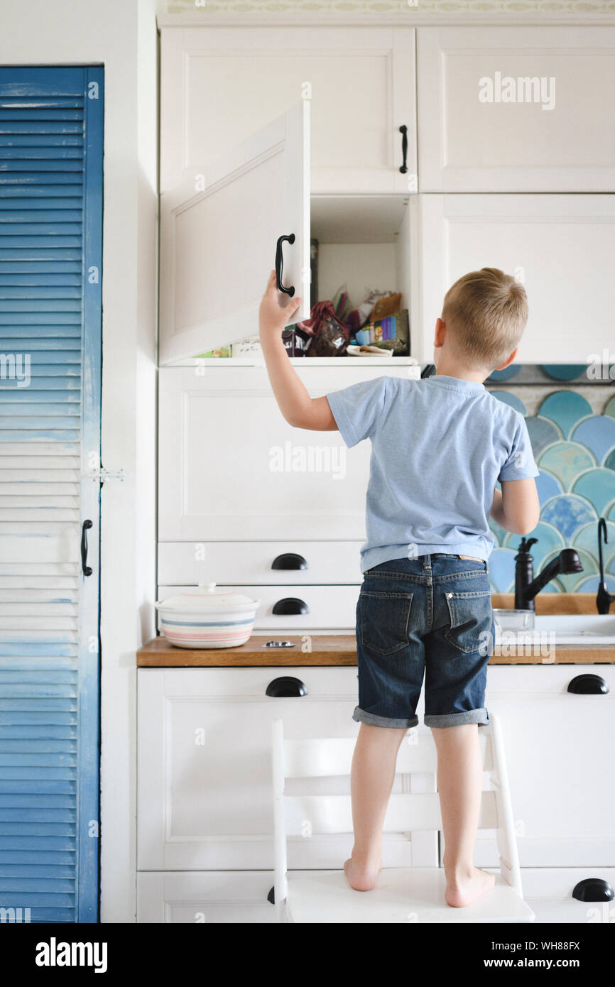 Back view of little boy standing on chair in the kitchen looking into cupboard Stock Photo