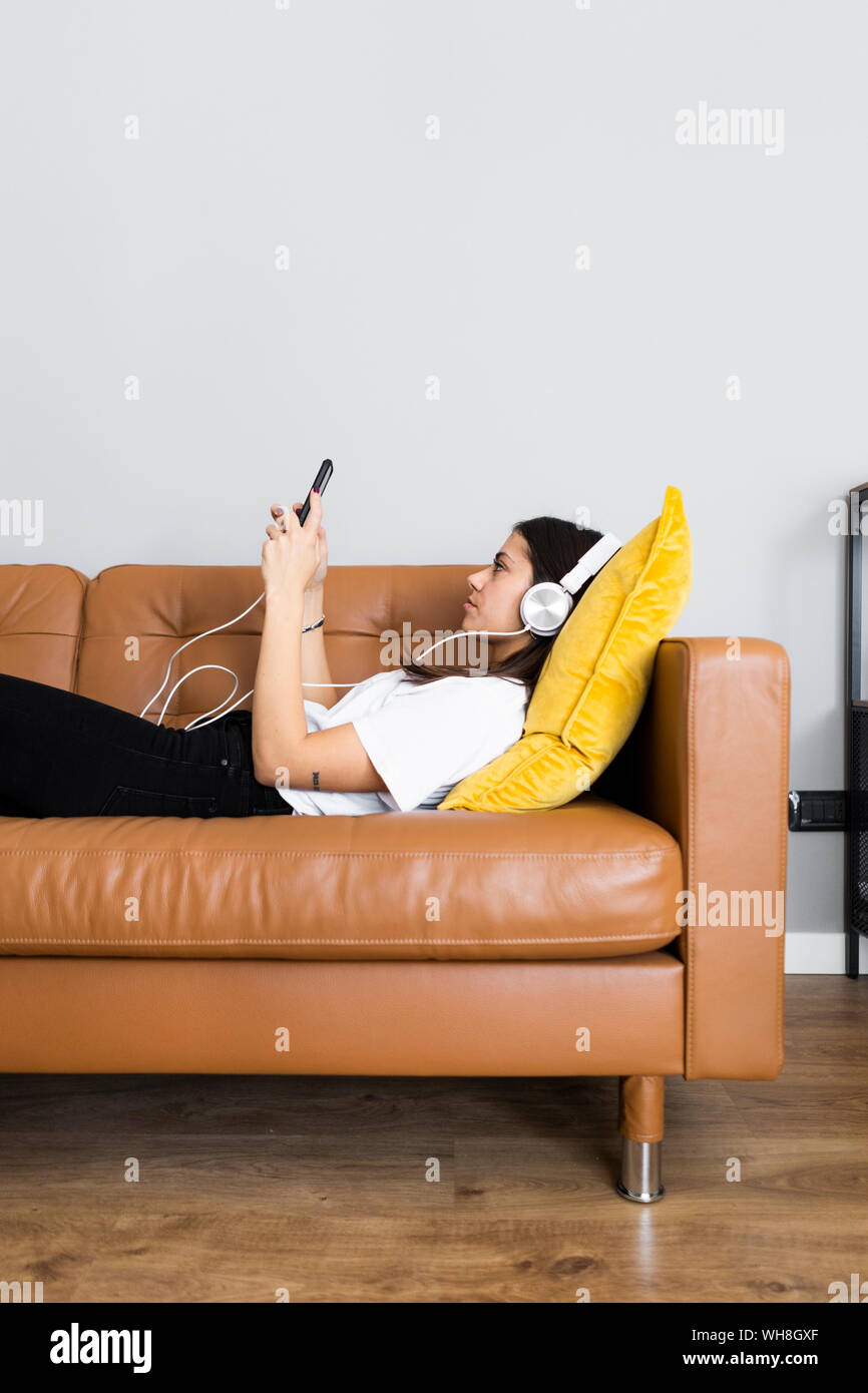Young woman lying on the couch at home with smartphone and headphones Stock Photo