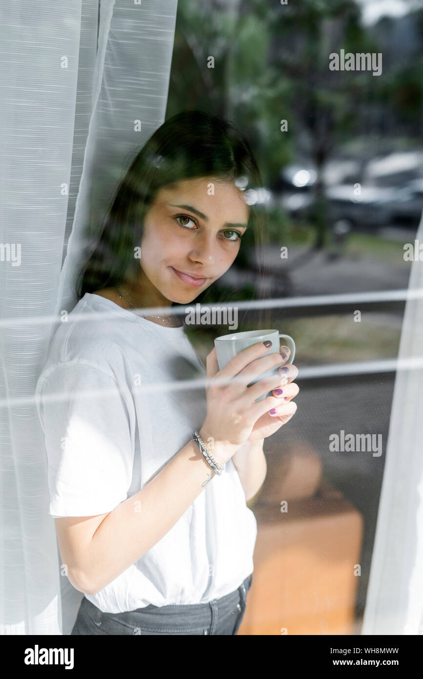 Portrait of young woman with cup of coffee behind windowpane Stock Photo