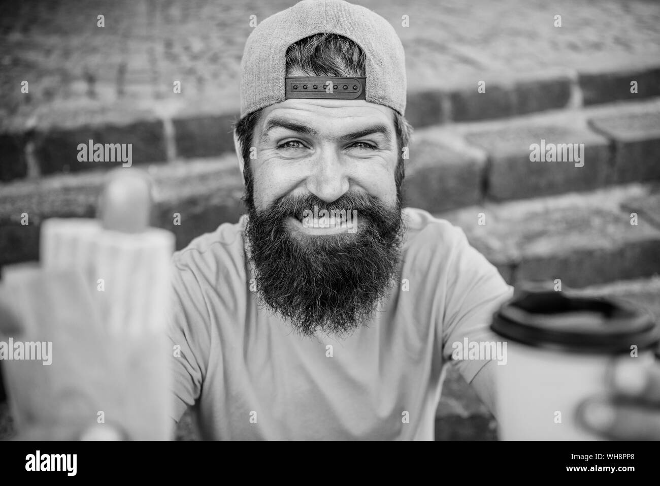 Man bearded eat tasty sausage and drink paper cup. Urban lifestyle nutrition. Junk food. Carefree hipster eat junk food while sit stairs. Snack for good mood. Guy eating hot dog. Street food concept. Stock Photo
