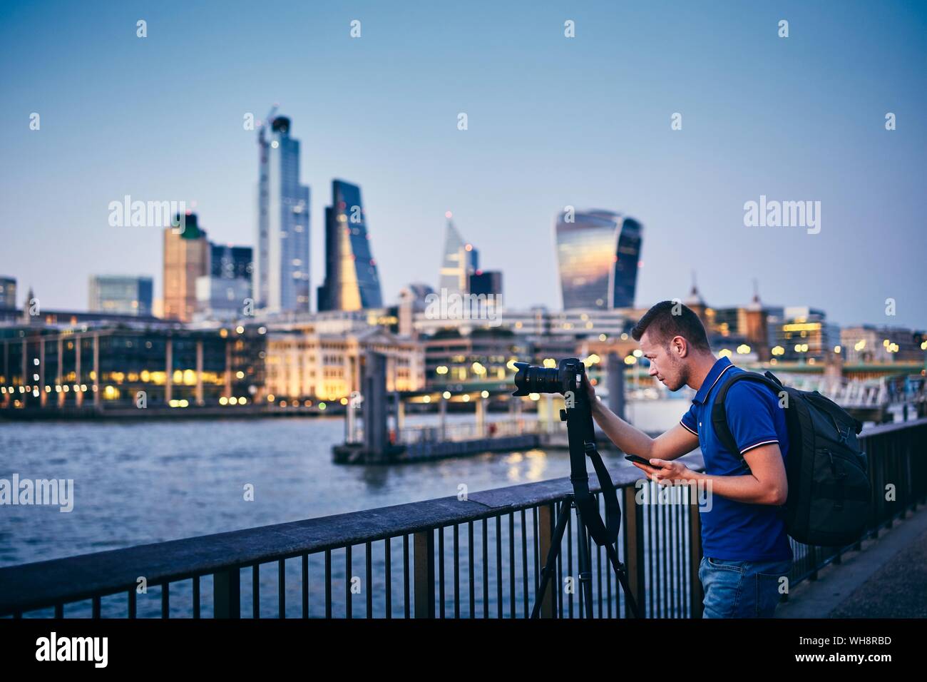 Young man photographing with tripod on embankment against urban skyline. Photographer holding smart phone with app. City life in London, United Kingdo Stock Photo