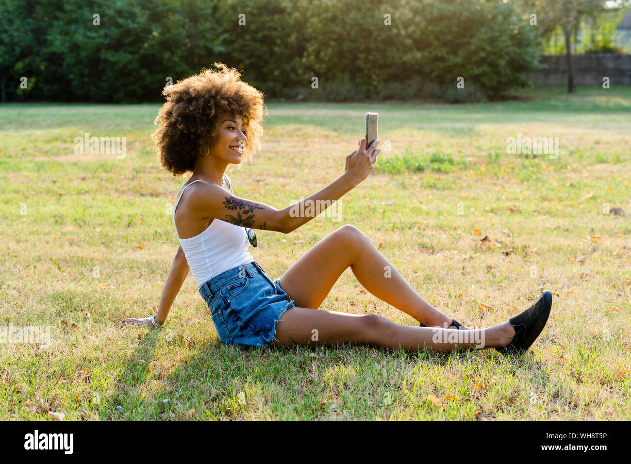 Tattooed young woman sitting on a meadow taking selfie with smartphone Stock Photo
