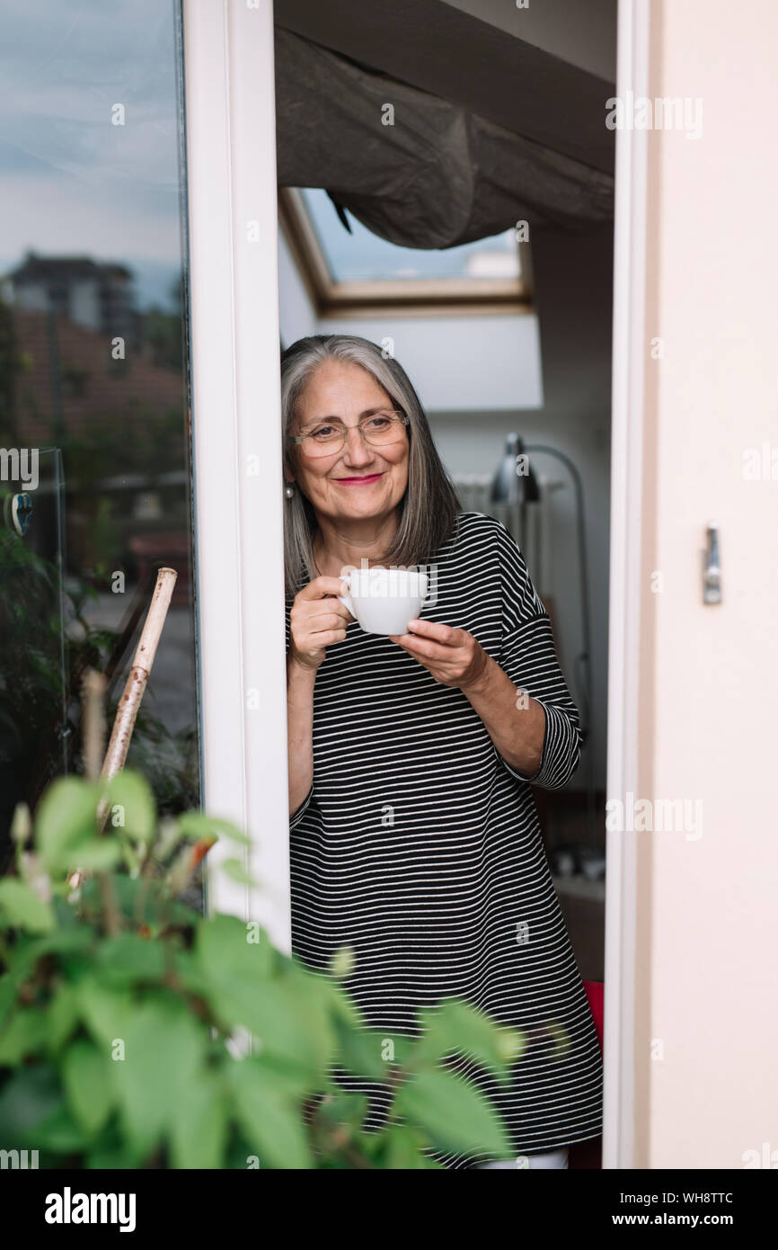 Portrait of happy senior woman leaning at opened balcony door with sup of coffee looking at distance Stock Photo
