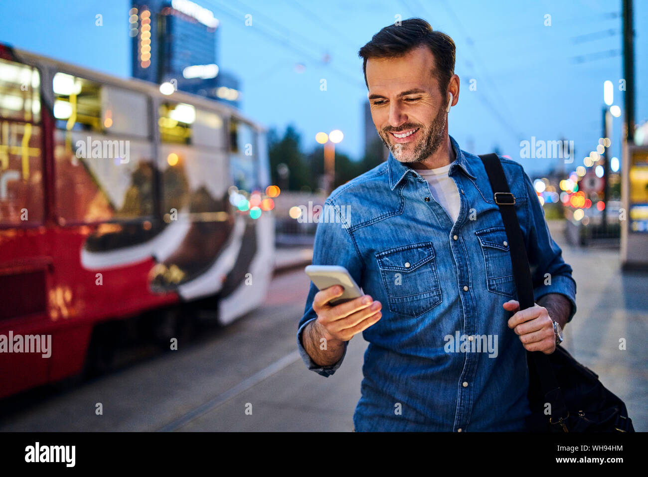Man using smartphone and listening to music while standing at tram stop in the evening Stock Photo