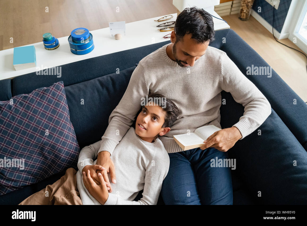 Father lying with son on couch in living room reading book Stock Photo