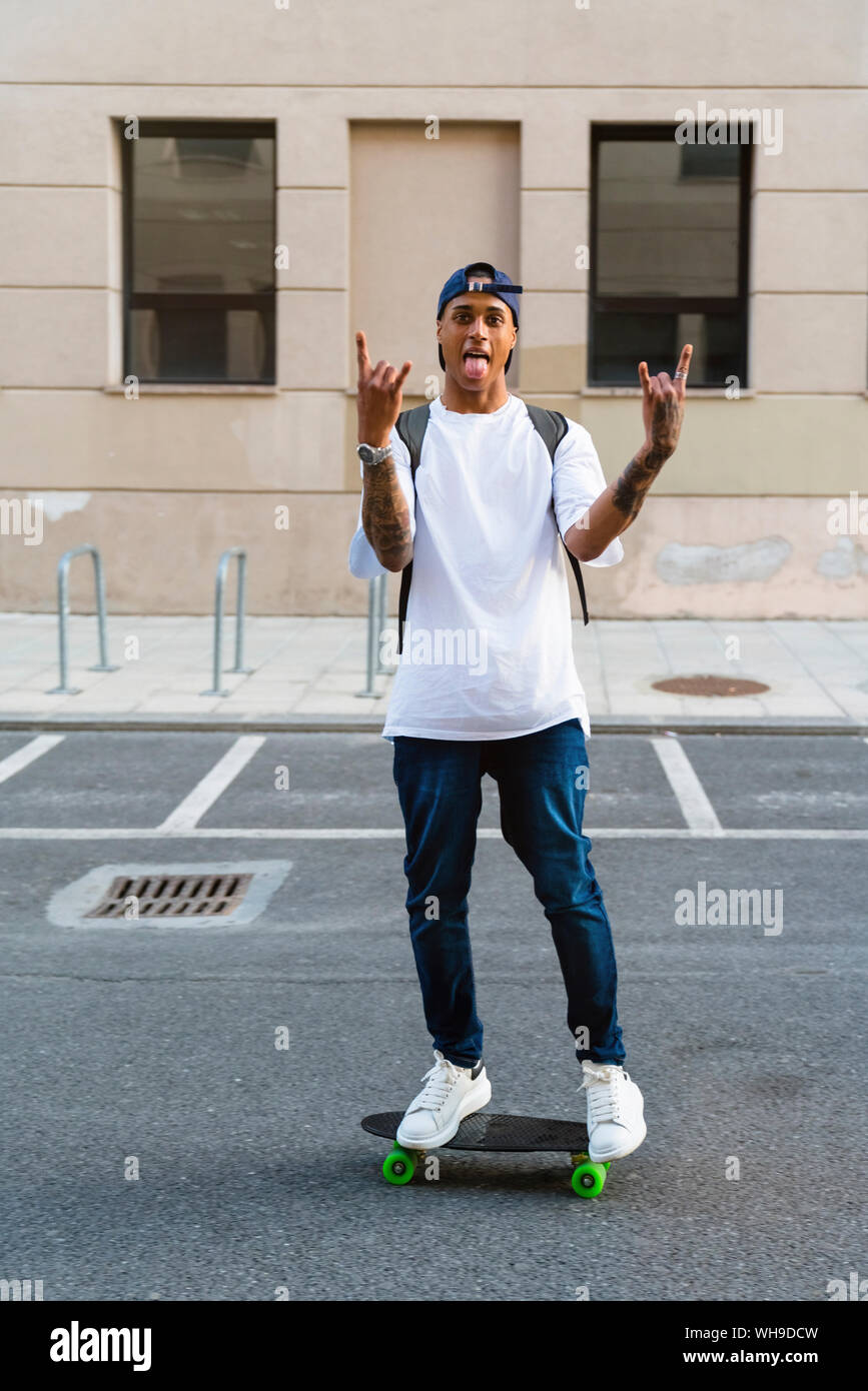 Tattooed young man standing on skateboard sticking out tongue and showing  Rock And Roll Sign Stock Photo - Alamy