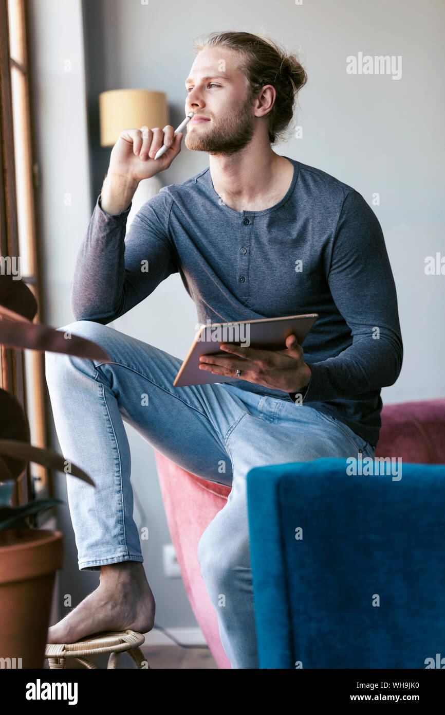 Pensive young man at home with a digital tablet looking out of a window Stock Photo