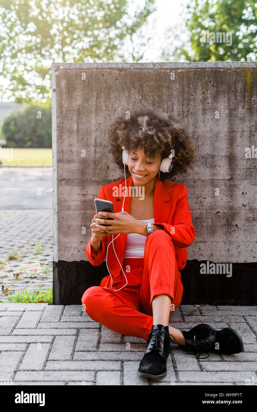 Portrait of smiling young woman with smartphone and headphones wearing fashionable red pantsuit Stock Photo