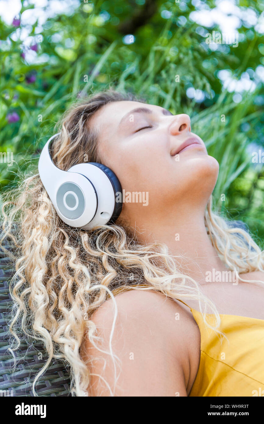 Smiling woman listening to music with closed eyes, lying on deckchair Stock Photo