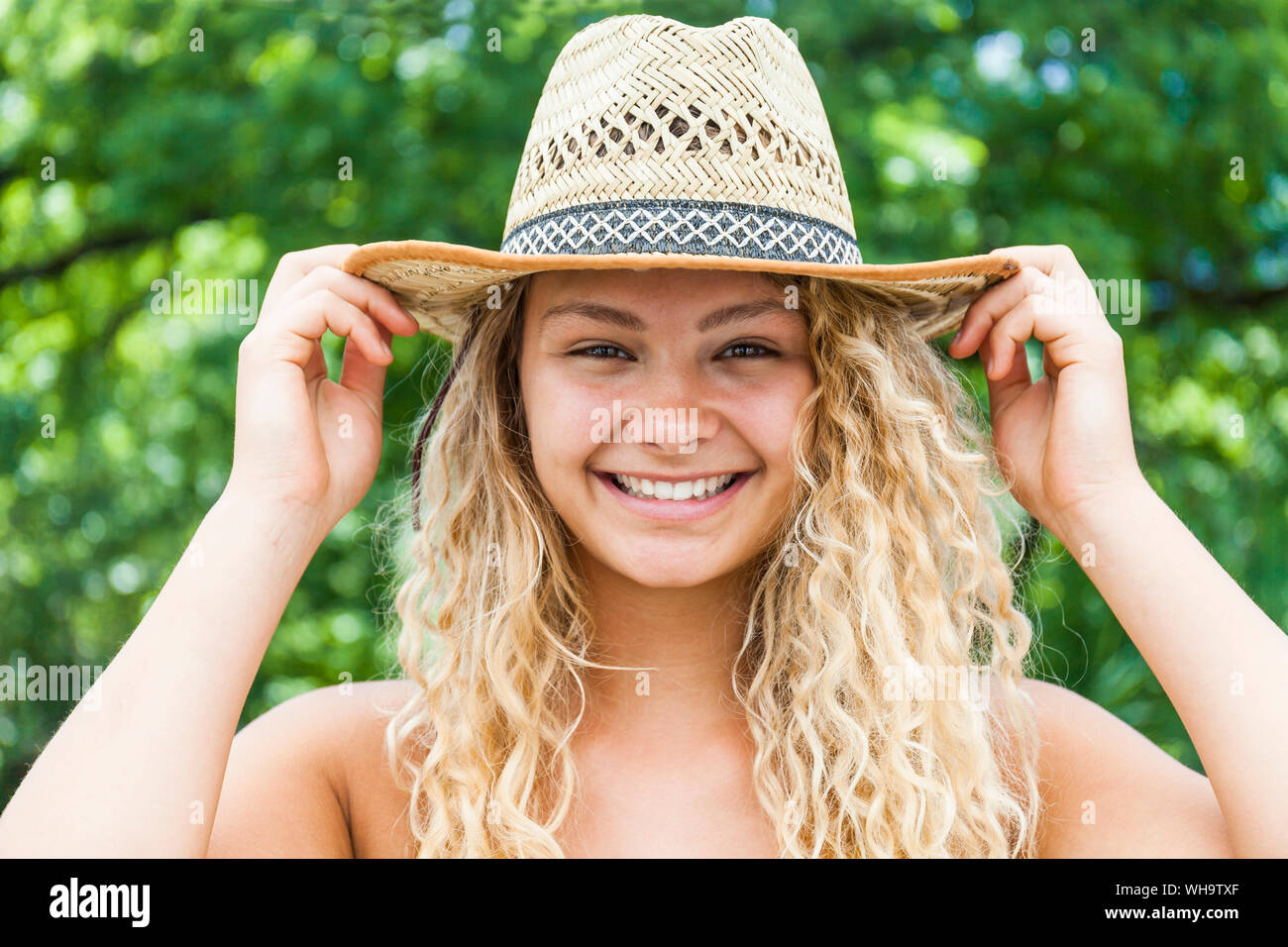 Portrait of smiling blond woman wearing straw hat, hands on hat Stock Photo