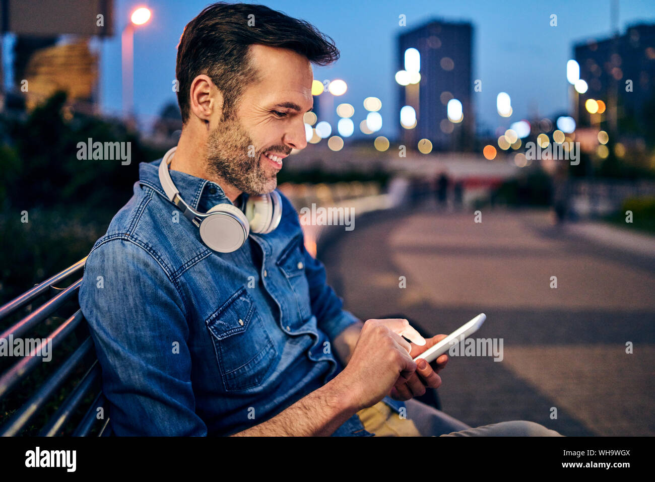 Smiling man using his smartphone while sitting on a bench in the evening Stock Photo
