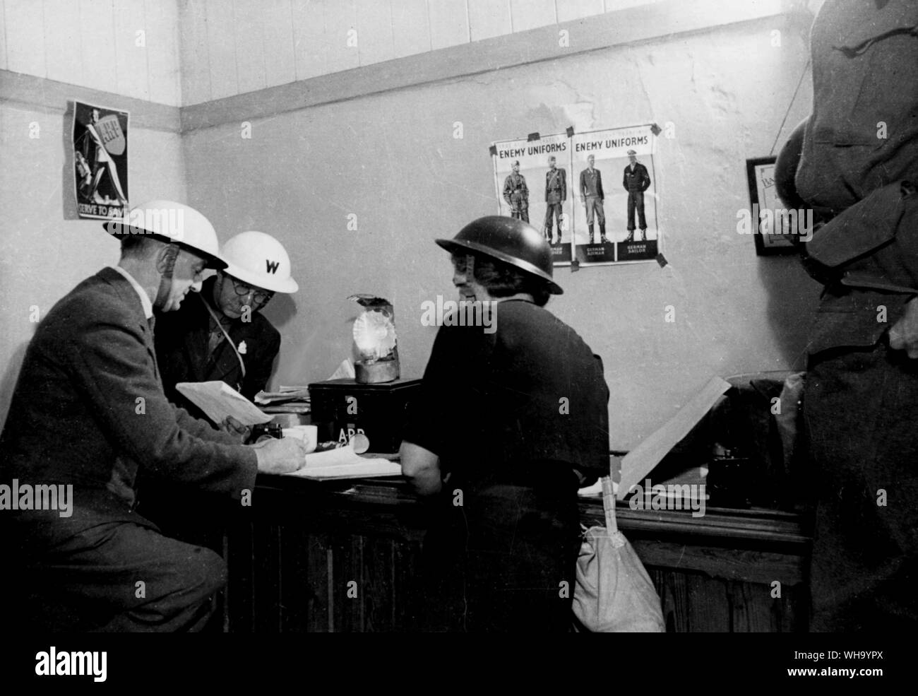 WW2: Invasion Village, like everyday city town hall and hamlet, throughout Britain, it's Warden's post. Head Warden Elliott, Warden Stern and Warden Nancy Roberts (l-r) work with the aid of an oil lamp. On the wall are posters giving identification of German Uniforms and another calling ARP workers. Stock Photo