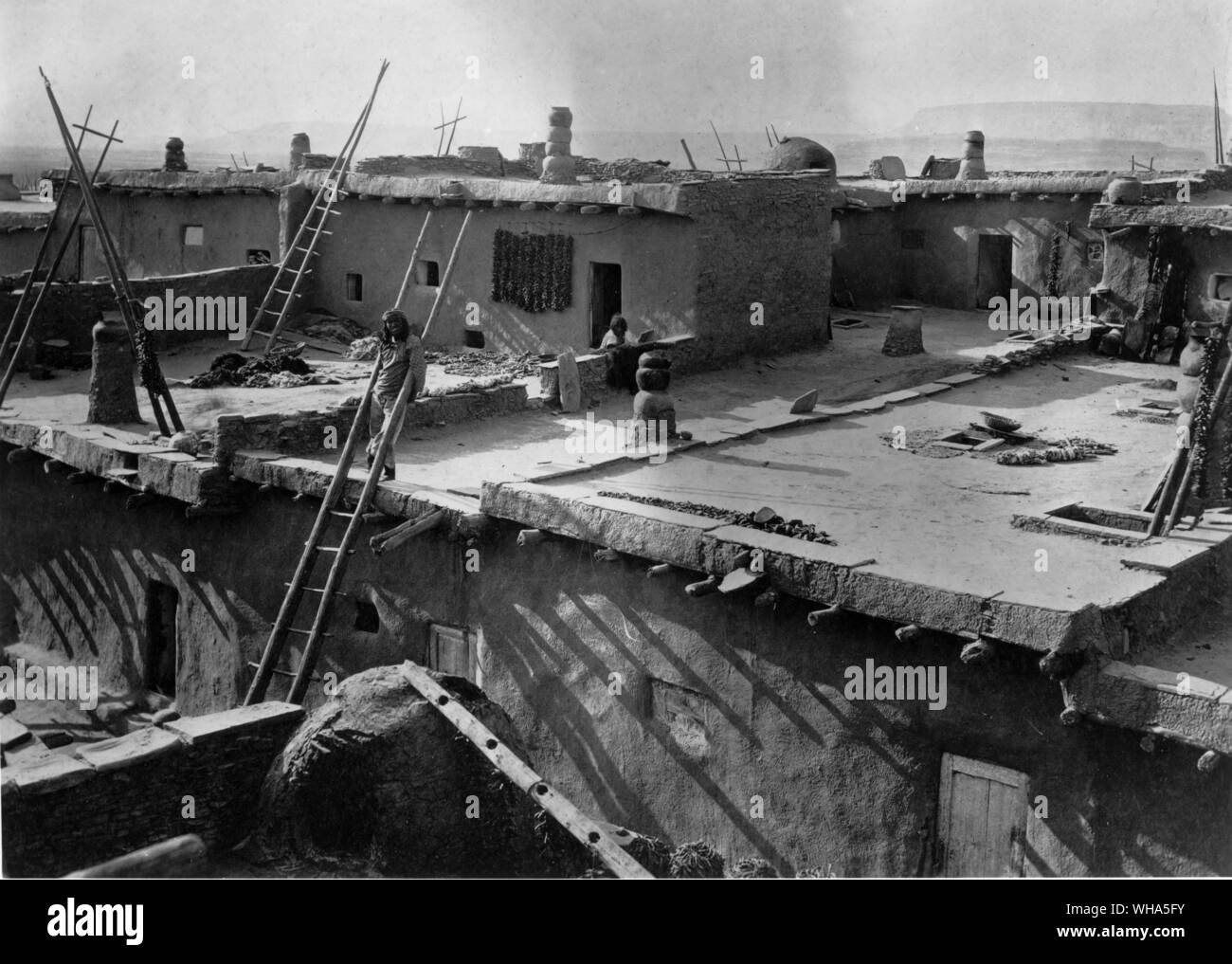 1879. Typical terraced pueblo type of architecture. Closeup shows how the rooftops of the lower levels were used as dooryards for the higher stories. Many of the household tasks such as drying fruits and vegetables were and are performed on these areas. The rectangular opening with projecting ladder in the right hand roof indicates that the room below is a ceremonial chamber. During ceremonies all entering and leaving is through the roof opening.Under normal circumstances the door on the ground level is used. The other rectangular openings in that particular roof are for the admittance of Stock Photo