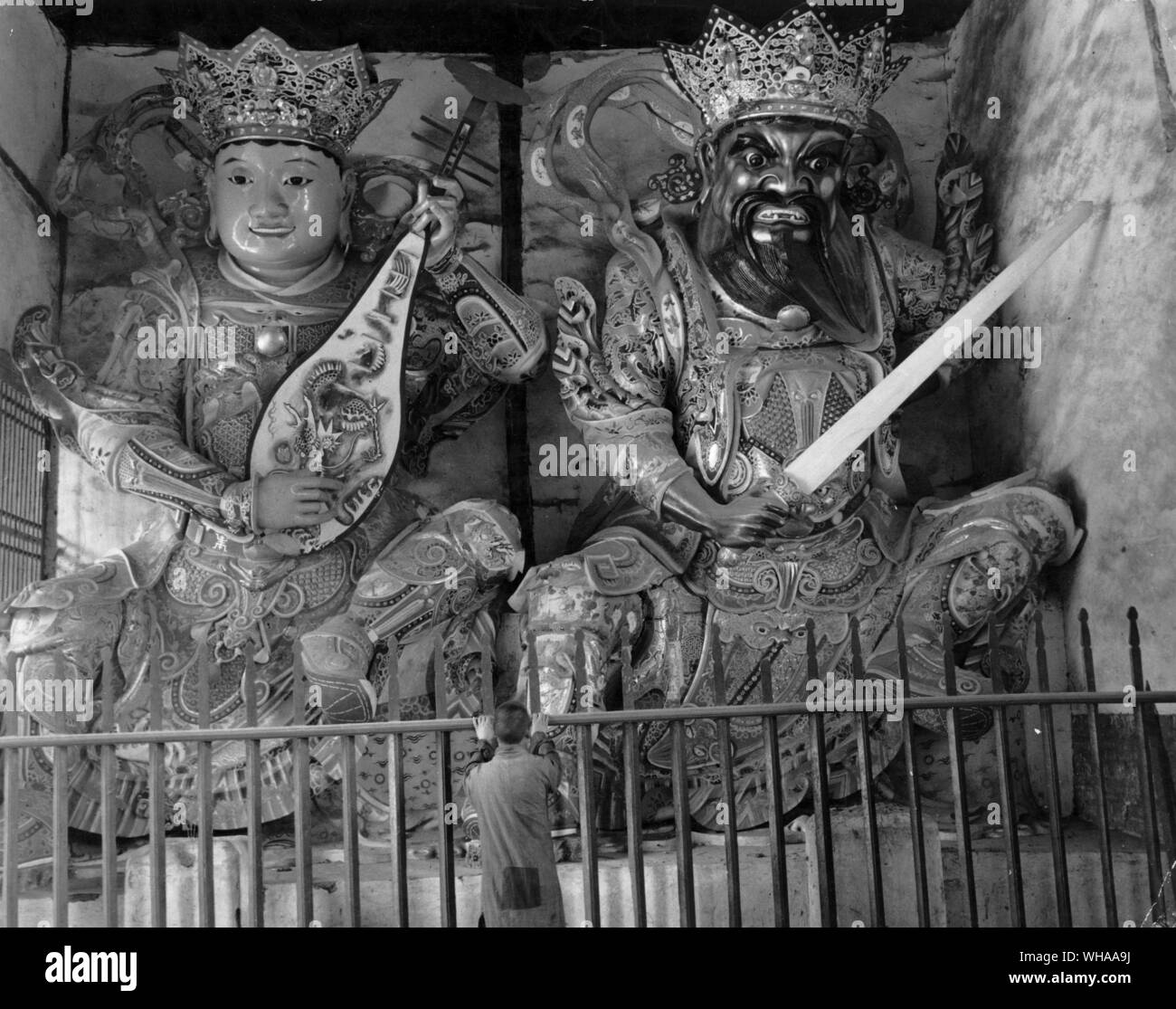 The yearly pilgrimages of the Chinese peoples to the Buddhist Temples where they worship Gautama Buddha, his idols and guardians, are like national holidays for them. Entire families young and old set out on foot for the long and tedious trek to the hills where the temples stand in lofty solitude. . Picture shows a Buddhist monk centre stands praying before the oversized carved and highly decorated crowned statues of the two Buddhistic symbols possibly portraying evil and good spirit Stock Photo