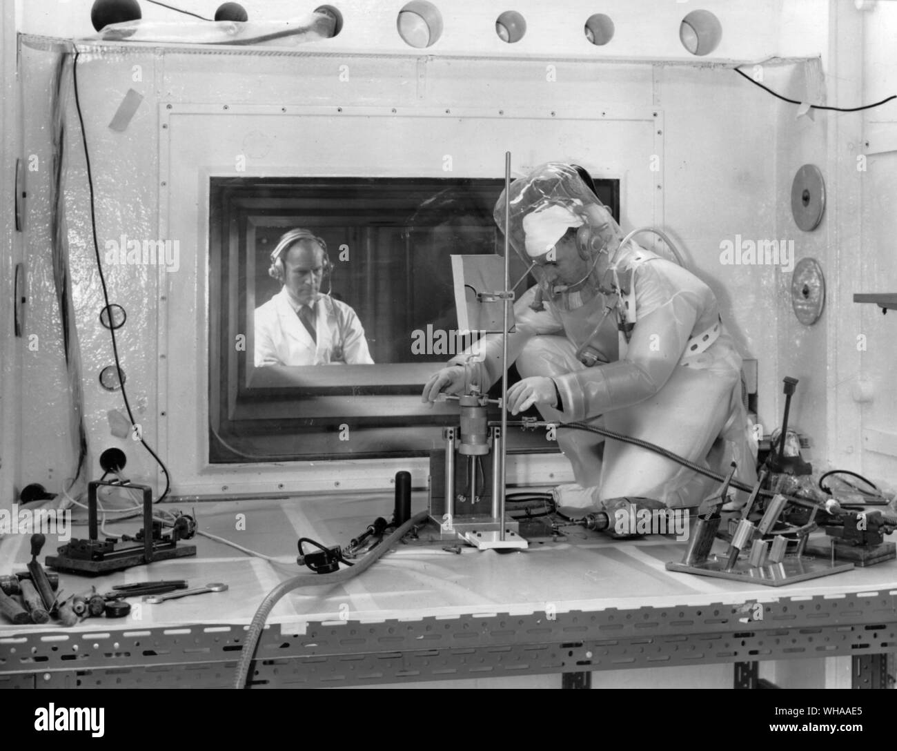 Research into the effects of radiation on materials is carried out in the High Activity Handling Building.. The work is done in cells which have protective walls of concrete 5ft 6 inches thick using remote manipulators and remotely controlled machines. Operations are watched through large windows also 5 ft 6 inches thick which are glass fronted tanks filled with zinc bromide solution. A maintenance mechanic in protective clothing adjusting equipment in a highly active cell. The work is being directed from behind the glass shielding window. . Harwell November 1962 Stock Photo