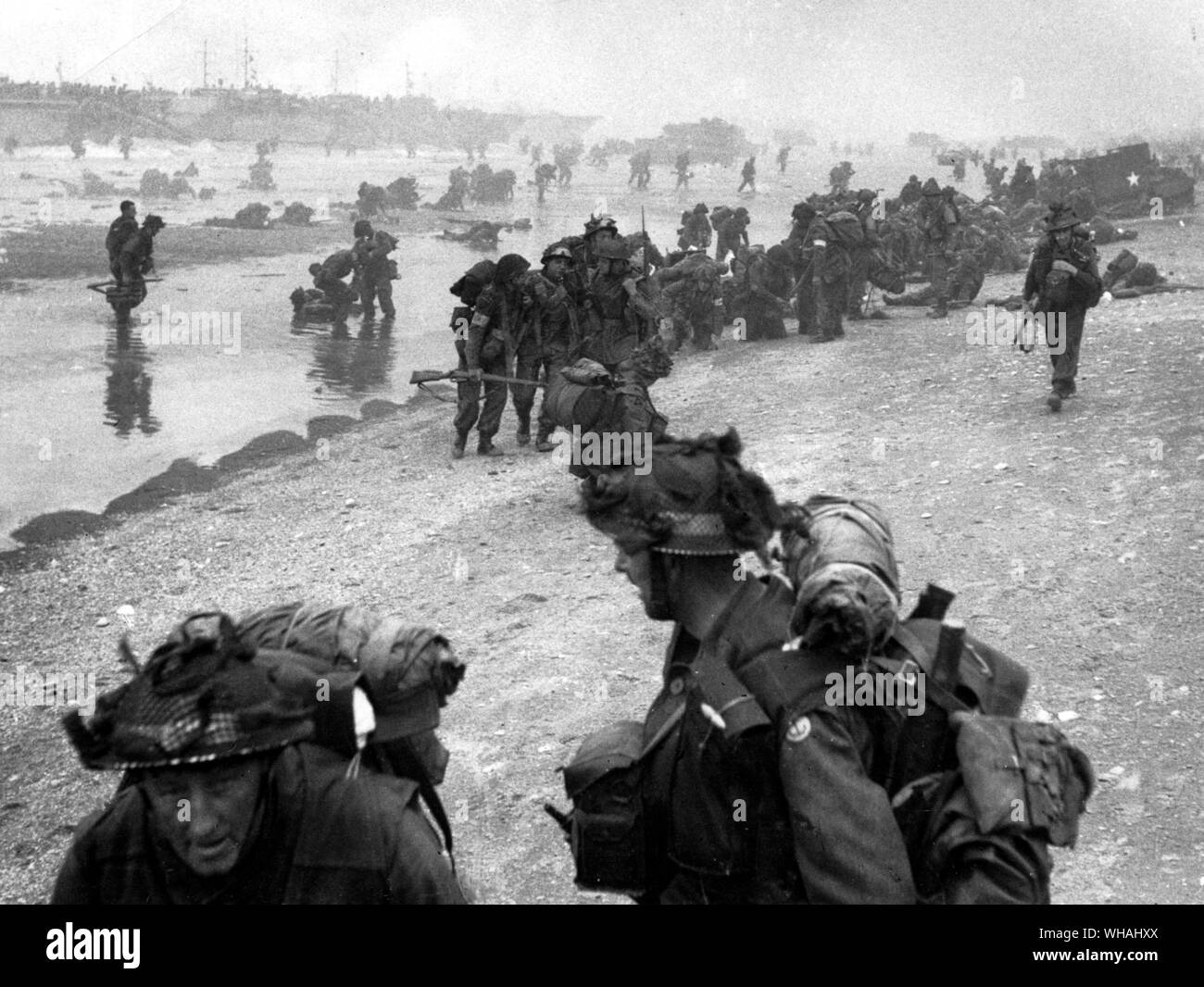 Troops assembling on beach from landing craft. Red Cross men attending to casualties. Broch Group? 13/18th Hussars. White Beach. 6th June 1944 D-Day. . WW2: Troops assembling on the beach from landing craft in the background. Red Cross men attending to casualties. Broch Group markings. 13/18th Hussars. White Beach, D day. 6th June 1944. Stock Photo