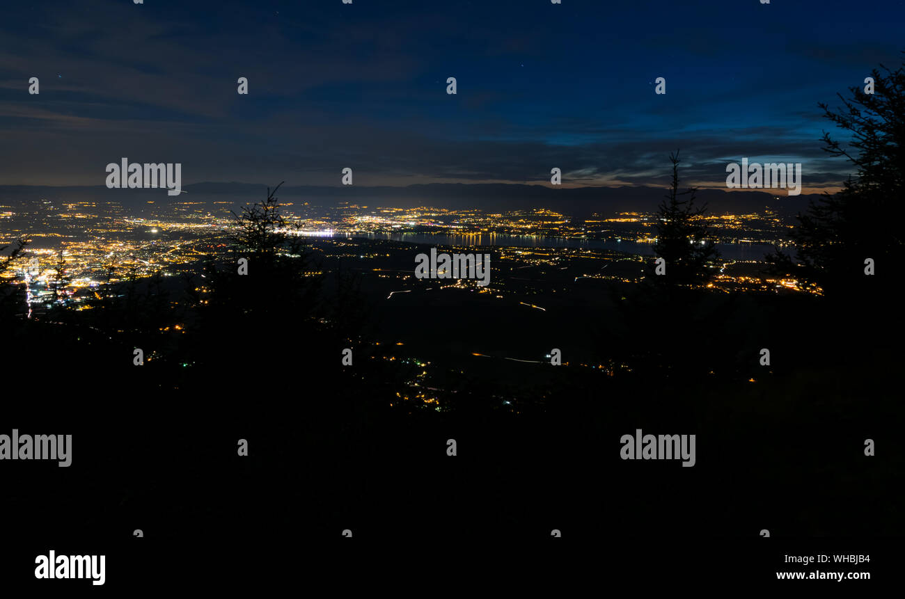 Evening top view of city lights, French Annemasse, Swiss Geneva,lake Geneva and picturesque sky with dark clouds after sunset,photo with long exposure. Stock Photo