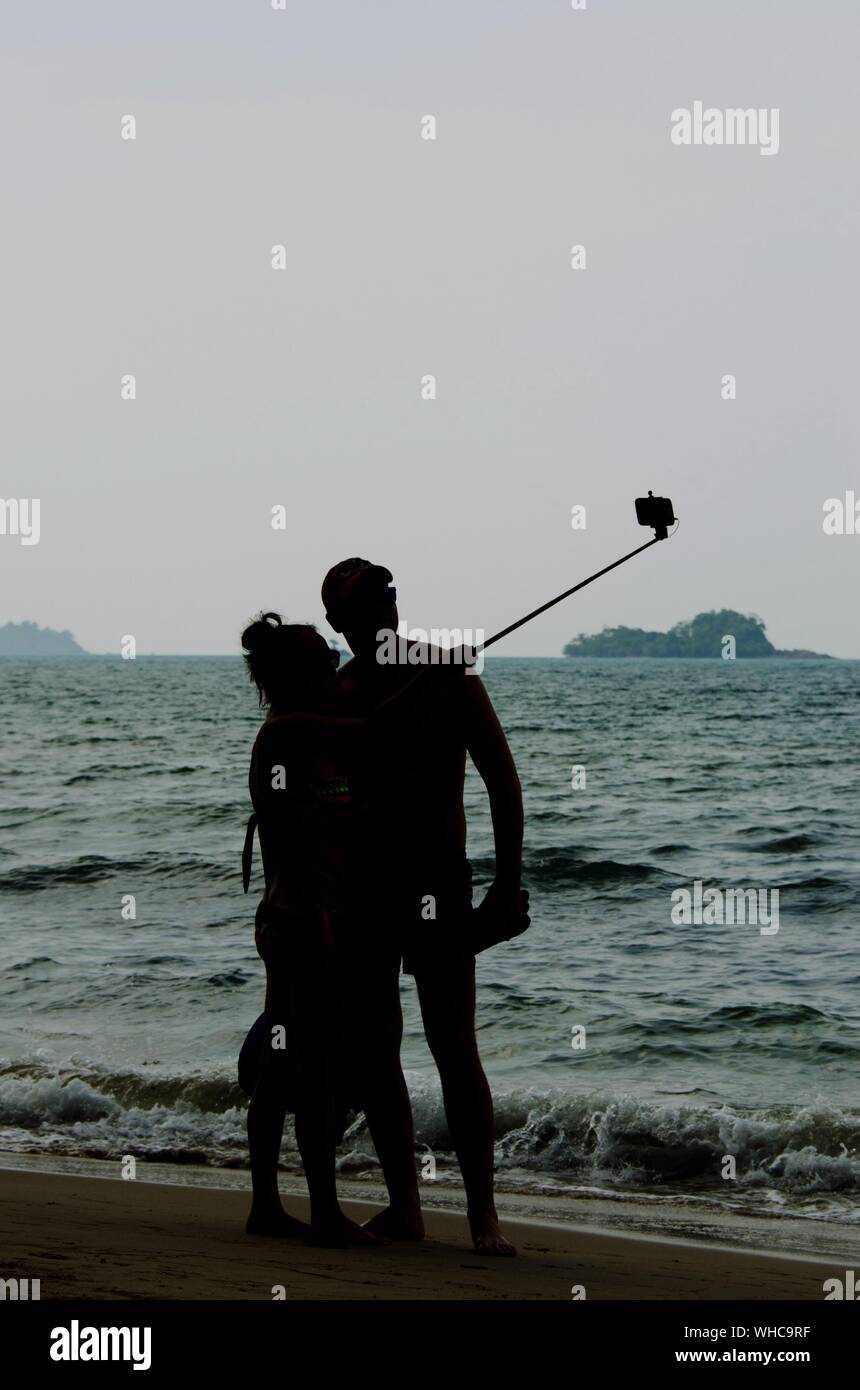 Silhouette Couple Clicking Selfie At Beach Against Sky Stock Photo