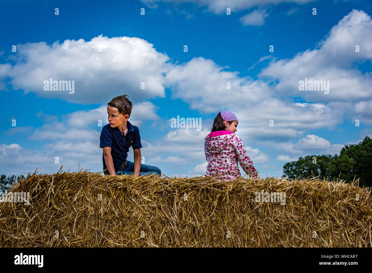 iconic picture of a lovely little boy and a  lovely little girl sitting on a hay stack against a beautiful blue sky with clouds Stock Photo